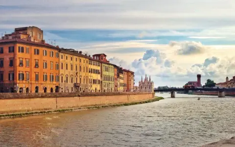 Edifici colorati lungo il fiume Arno a Pisa, con un ponte e nuvole suggestive nel cielo. Un'immagine che cattura la bellezza di Pisa e dintorni.