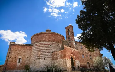 L'abbazia di San Galgano, con la sua rotonda in mattoni a fasce bianche e rosse, si staglia sotto un cielo azzurro con nuvole bianche.