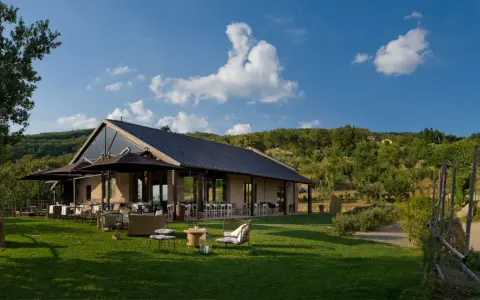 Il podere di pomaio, con tavoli all'aperto e un prato verde, si estende su una collina alberata sotto un cielo azzurro.