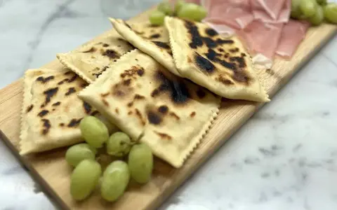 Tortelli alla lastra casentino, dorati e leggermente bruciacchiati, serviti con uva fresca e affettati di prosciutto su un tagliere di legno.