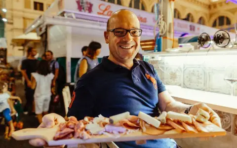 Uomo sorridente al mercato delle vettovaglie Livorno, offre un tagliere di salumi e formaggi.