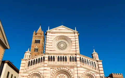 Il Duomo di Grosseto, con la sua facciata a strisce bianche e rosa e il campanile in mattoni, si staglia contro un cielo azzurro intenso.
