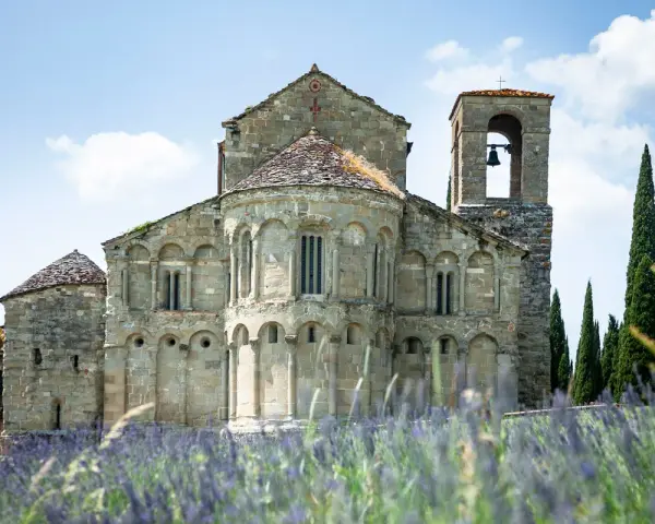 La Pieve di Romena, un'antica chiesa in pietra con campanile, si erge maestosa tra cipressi e un campo fiorito di lavanda sotto un cielo azzurro.
