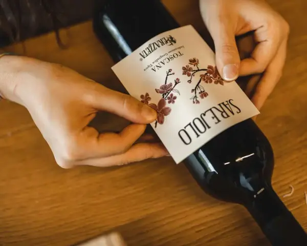 Mani che applicano un'etichetta su una bottiglia di vino. L'etichetta indica "Toscana", la regione di origine del vino Sangiovese.