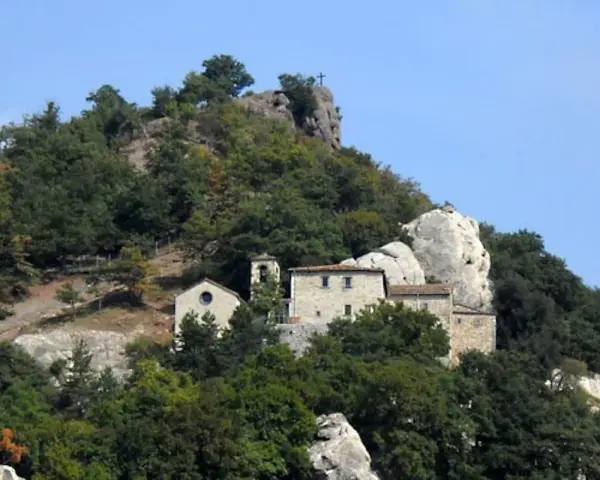 Antico eremo di Cerbaiolo, un complesso di edifici in pietra arroccato su un colle boscoso, con una croce sulla cima di una roccia.