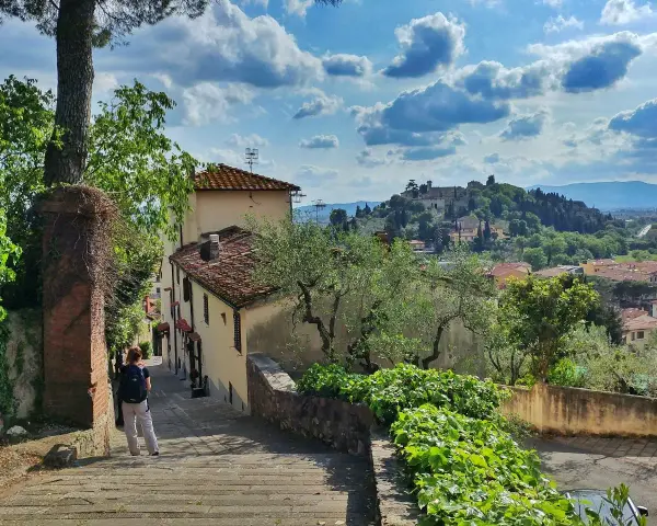 Una persona scende una scalinata in pietra, con case antiche e vegetazione rigogliosa. In lontananza, il **monte Morello** domina il paesaggio collinare.