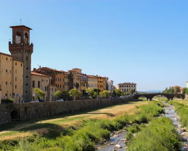Pescia: cosa vedere? Un fiume serpeggia tra case colorate e un'antica torre con orologio, sotto un cielo azzurro.