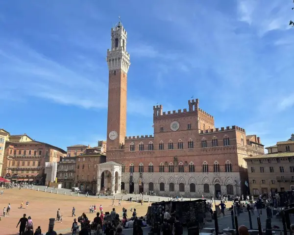 La Piazza del Campo di Siena, con la Torre del Mangia che domina il cielo azzurro, brulica di gente che passeggia e si gode il sole.