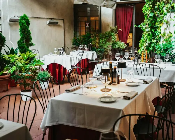 Cortile di un ristorante, tavoli apparecchiati con tovaglie bianche e rosse, piante verdi e sedie nere. Un invito a mangia e bevi santo spirito.