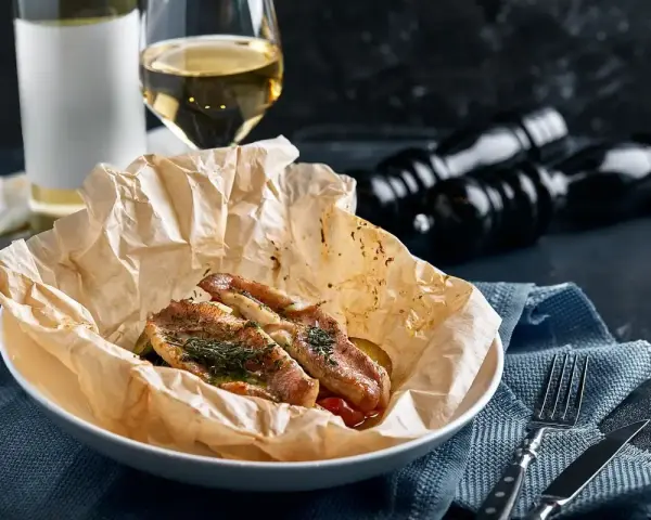 Deliziosa trota puccini cotta al forno, servita su carta da forno con erbe aromatiche. Accanto, un bicchiere di vino bianco e macinapepe.
