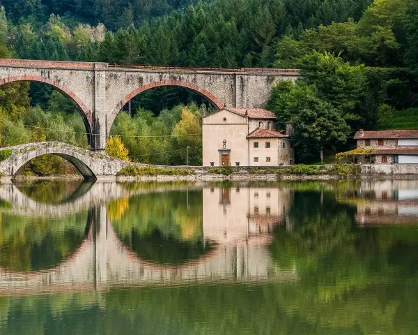 Pieve Fosciana: due ponti, uno antico e uno moderno, si specchiano in un lago calmo, con edifici storici e una foresta sullo sfondo.