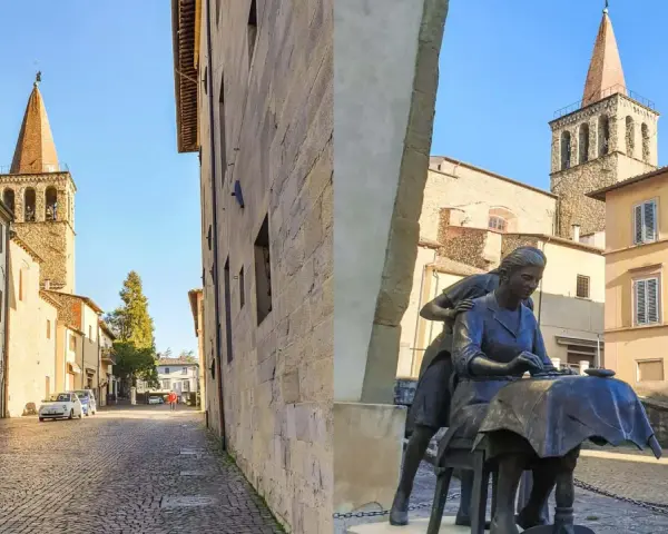 Statua di donne al lavoro, con campanili e palazzi storici sullo sfondo. Un assaggio di cosa vedere a Sansepolcro.