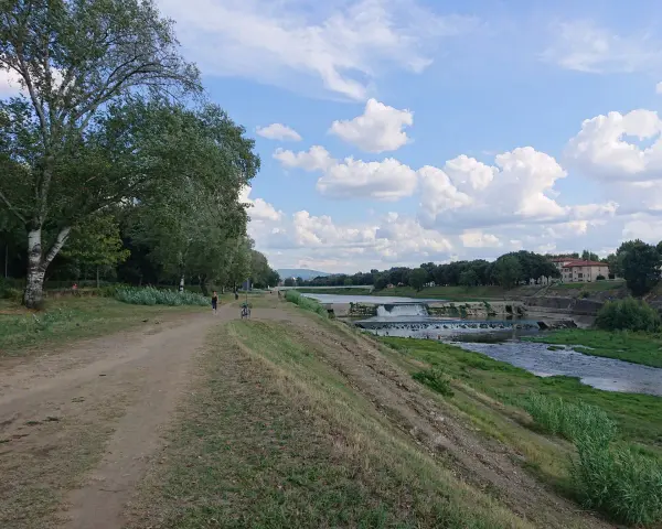 Sentiero lungo il fiume con alberi e il masso della Gonfolina in lontananza. Cielo azzurro con nuvole bianche.
