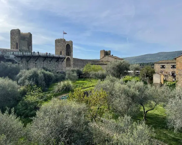 Le mura di Monteriggioni, Siena, dominano un paesaggio toscano con ulivi e colline. Un ponte pedonale attraversa la fortificazione.