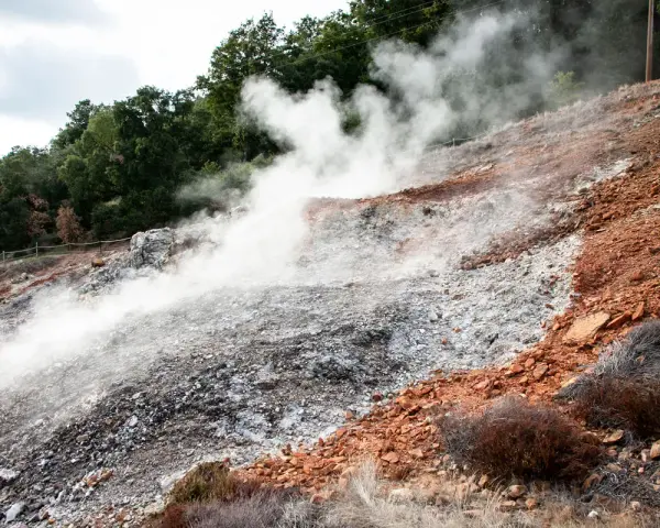 Fumarole attive nel Parco delle Biancane, con vapore che sale da un terreno roccioso e argilloso, circondato da vegetazione e alberi.