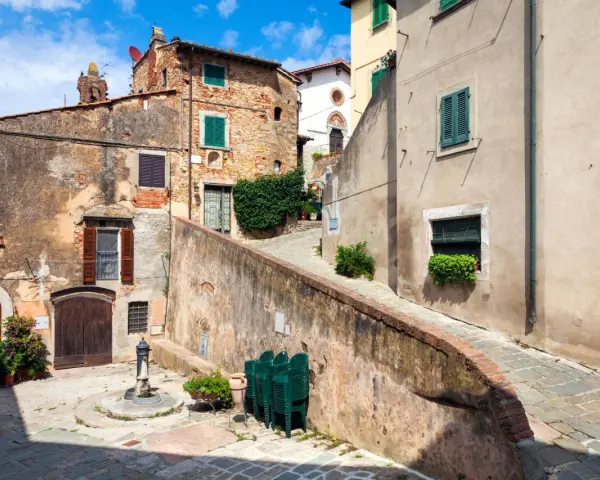 Un vicolo pittoresco a Castagneto Carducci, con case antiche, un fontanello e sedie verdi impilate.