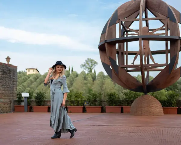 Donna con cappello nero ammira l'Uomo Vitruviano in una sfera di legno, un'opera iconica da vedere a Vinci, con colline verdi sullo sfondo.