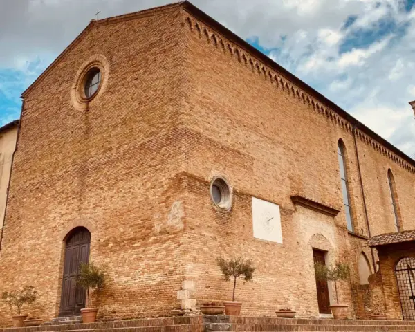 La **chiesa di Sant'Agostino** a San Gimignano, con la sua facciata in mattoni rossi e il campanile svettante, si staglia contro un cielo azzurro e nuvoloso.