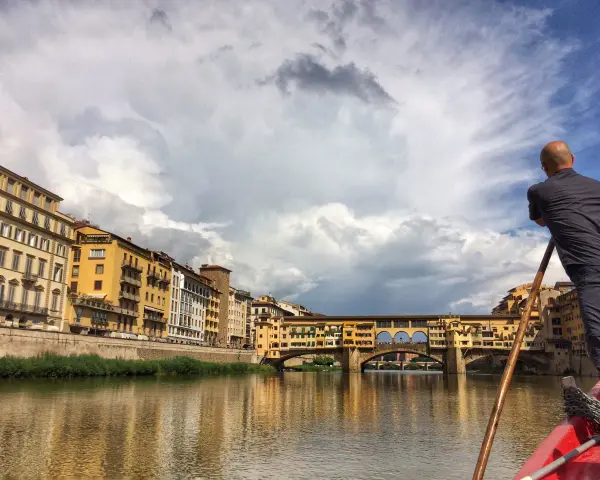 Un uomo su una barca a remi naviga lungo l'Arno, ammirando Ponte Vecchio e gli edifici storici di Firenze.
