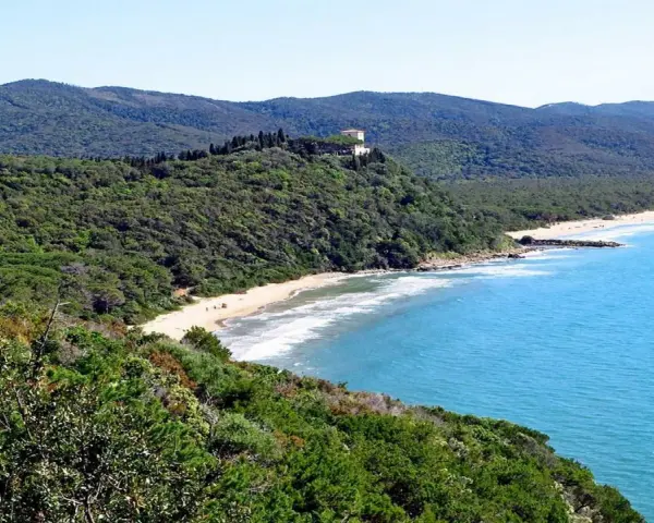 Vista panoramica delle bandite di Scarlino, con una spiaggia dorata bagnata da un mare azzurro e una villa in cima a una collina verde.