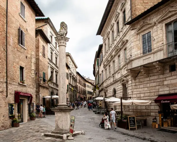 A stone column with a lion statue stands in a cobblestone street lined with historic buildings and shops.