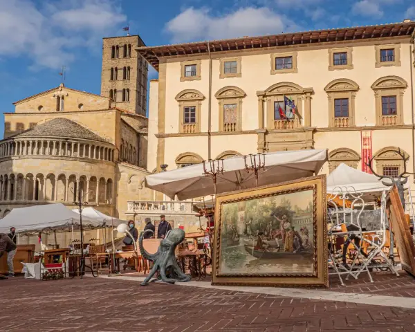 Mercatino dell'antiquariato in piazza grande, con opere d'arte, sculture e mobili antichi esposti sotto il sole.