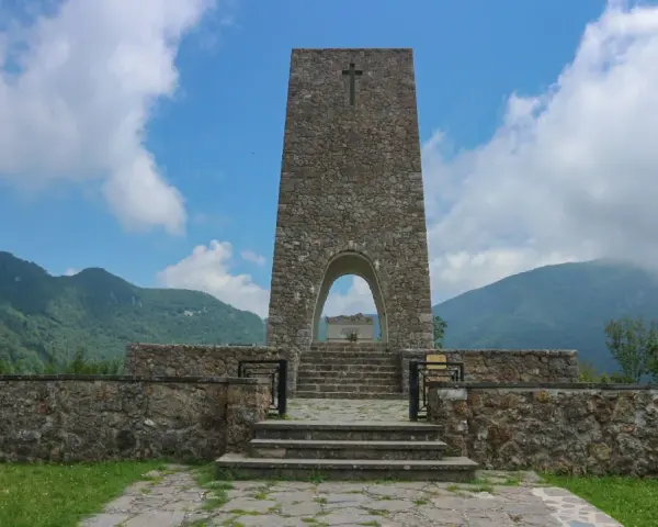 Monumento a Sant'Anna di Stazzema, cosa vedere: un imponente torrione in pietra con arco, che custodisce una tomba, circondato da mura e montagne.