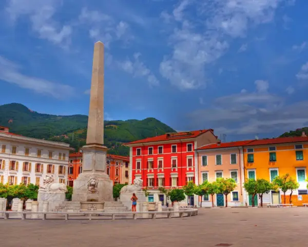 Piazza con obelisco e leoni, edifici colorati e montagne sullo sfondo. Un'idea di cosa vedere a Marina di Massa.