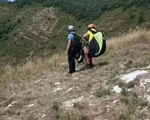 Due parapendisti si preparano al decollo sulla **Valle del Serchio**, con un paesaggio verdeggiante alle spalle.