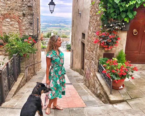 Una donna con un cane passeggia in un vicolo fiorito di Civitella Val di Chiana, con una vista panoramica sullo sfondo.