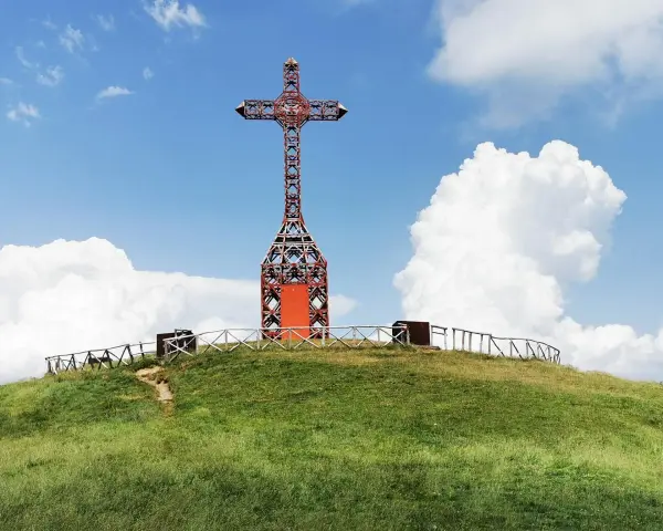 Croce metallica rossa sulla cima di una collina erbosa, con un sentiero che sale. Il **Pratomagno Arezzo** domina il paesaggio sotto un cielo azzurro con nuvole bianche.
