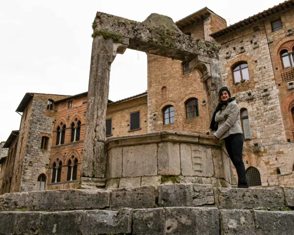 Donna sorride vicino a un antico pozzo a San Gimignano, con palazzi medievali sullo sfondo.