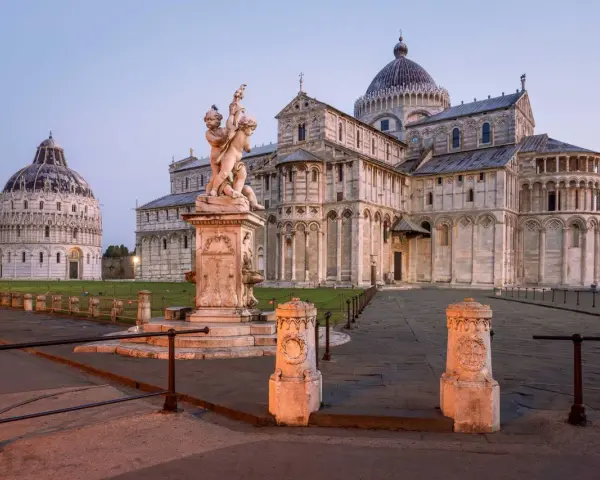 Il **Battistero di Pisa** e il Duomo illuminati dalla luce dell'alba. Una fontana con putti al centro della piazza.