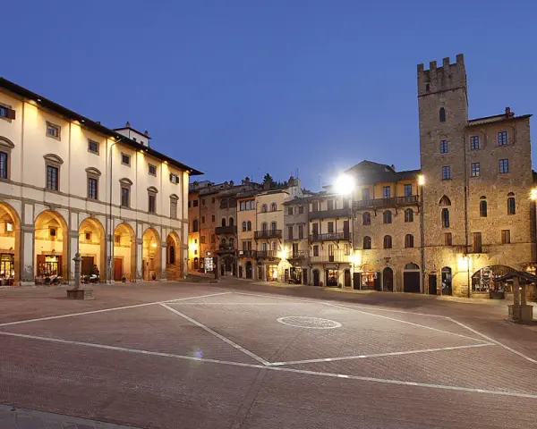 Piazza illuminata di Arezzo al crepuscolo, con portici, negozi e una torre medievale. Qui la vita è bella.