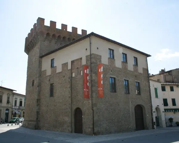 Il cassero di Montevarchi, un'antica torre con merlature, si erge imponente accanto a edifici storici, con bandiere rosse che adornano la facciata.