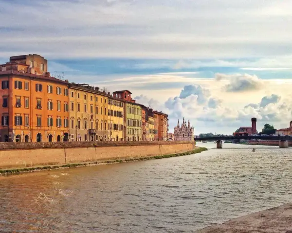 Edifici colorati lungo il fiume Arno a Pisa, con un ponte e nuvole suggestive nel cielo. Un'immagine che cattura la bellezza di Pisa e dintorni.