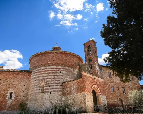 L'abbazia di San Galgano, con la sua rotonda in mattoni a fasce bianche e rosse, si staglia sotto un cielo azzurro con nuvole bianche.