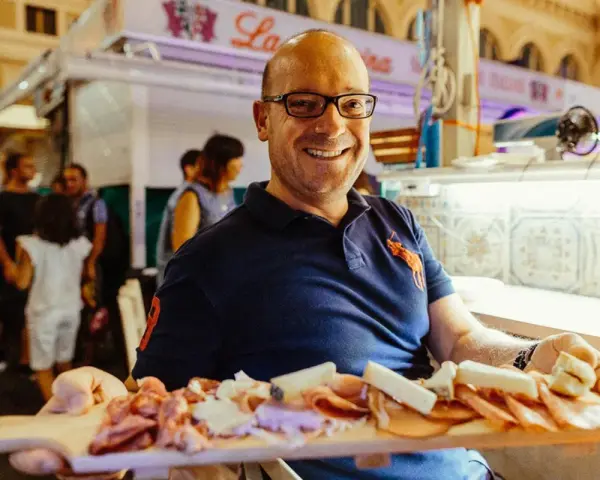 Uomo sorridente al mercato delle vettovaglie Livorno, offre un tagliere di salumi e formaggi.