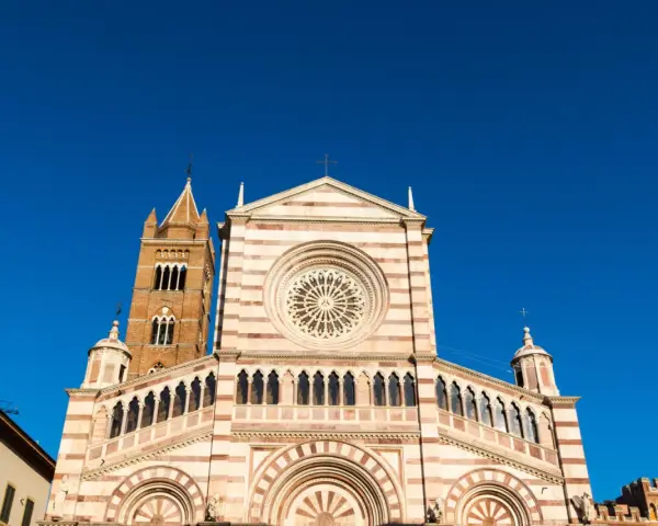 Il Duomo di Grosseto, con la sua facciata a strisce bianche e rosa e il campanile in mattoni, si staglia contro un cielo azzurro intenso.