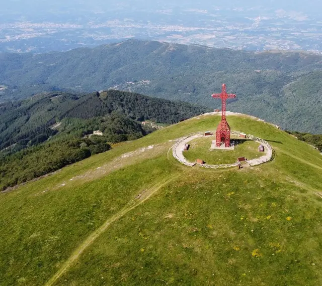 La croce del Pratomagno domina un paesaggio verdeggiante, con montagne boscose e una città in lontananza.
