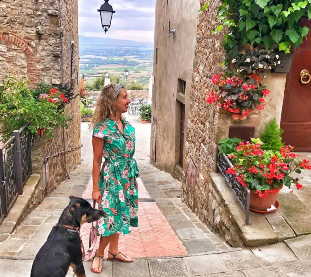 Una donna con un cane passeggia in un vicolo fiorito di Civitella Val di Chiana, con una vista panoramica sullo sfondo.