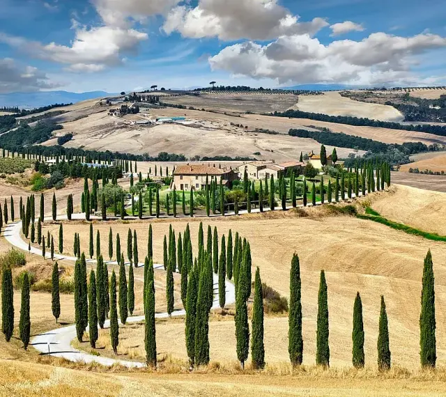 Un casale nel Val d'Orcia, circondato da cipressi e dolci colline dorate sotto un cielo azzurro con nuvole bianche.