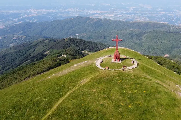 La croce del Pratomagno domina un paesaggio verdeggiante, con montagne boscose e una città in lontananza.