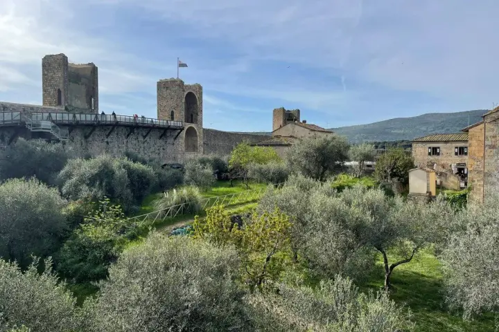 Le mura di Monteriggioni, Siena, dominano un paesaggio toscano con ulivi e colline. Un ponte pedonale attraversa la fortificazione.