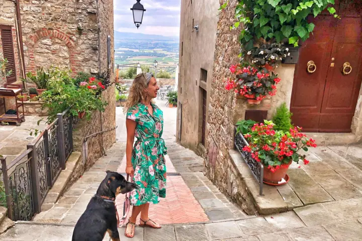 Una donna con un cane passeggia in un vicolo fiorito di Civitella Val di Chiana, con una vista panoramica sullo sfondo.