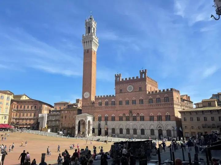 La Piazza del Campo di Siena, con la Torre del Mangia che domina il cielo azzurro, brulica di gente che passeggia e si gode il sole.