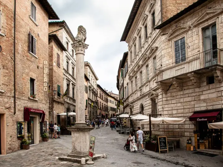 A stone column with a lion statue stands in a cobblestone street lined with historic buildings and shops.