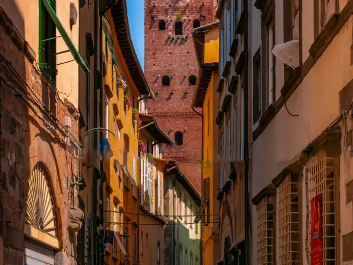 Un vicolo stretto a Lucca con edifici colorati e la torre Guinigi che svetta in lontananza.