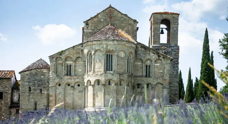 La Pieve di Romena, un'antica chiesa in pietra con campanile, si erge maestosa tra cipressi e un campo fiorito di lavanda sotto un cielo azzurro.