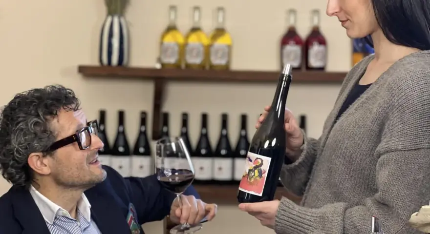 Uomo con occhiali e bicchiere di vino rosso, donna presenta bottiglia. Degustazione vini Arezzo in cantina.